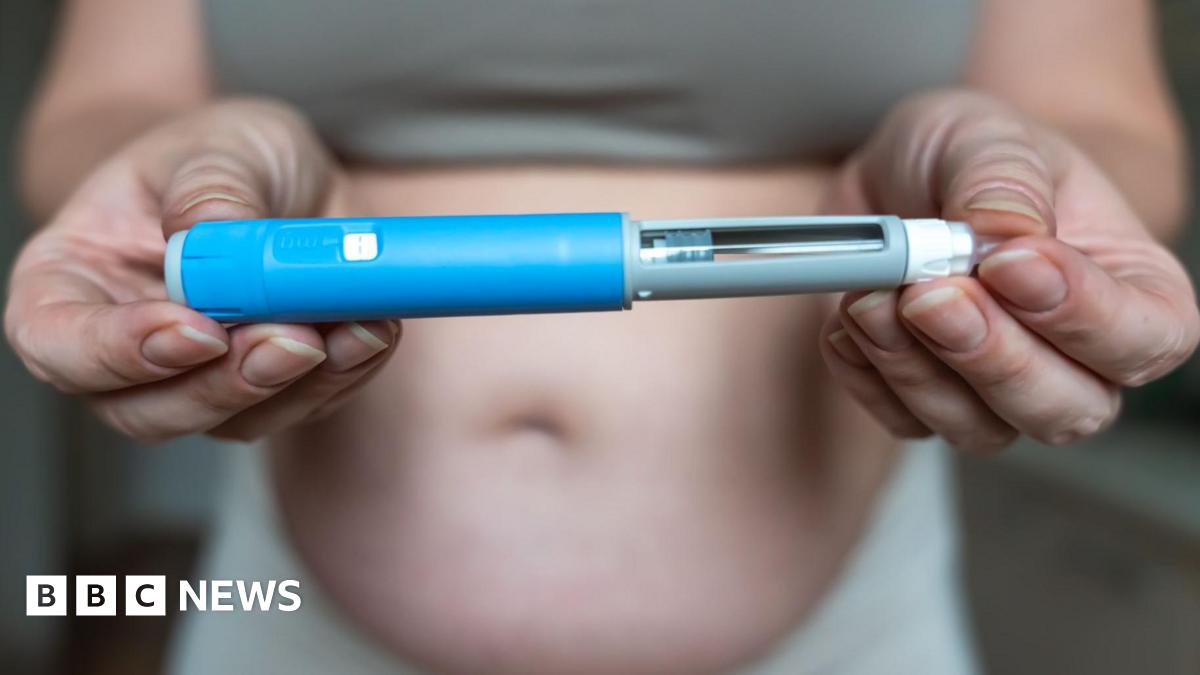 A person holding an injector pen for a weight-loss drug - the pen is in focus at the foreground of the image, held at either end by both hands of the person. To the left, the pen has a blue outer shell, with it then tapering to the right into a grey section, with a see-through strip, to the sharp point of the needle. The person in the background appears to be a woman, we only see their middle third. They are wearing a top that cuts off at the belly, showing the person is overweight.