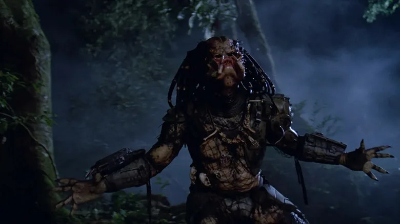 The Predator screaming with his hands open wide in the dark jungle