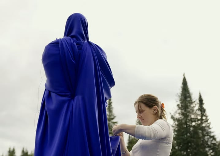 A woman adjusts a large, deep blue fabric draped over a tall, human-like figure outdoors, with evergreen trees and an overcast sky in the background.