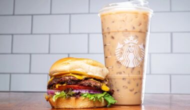 A cheeseburger with lettuce, tomato, onions, and pickles sits next to a large iced coffee in a clear Starbucks cup on a wooden surface.