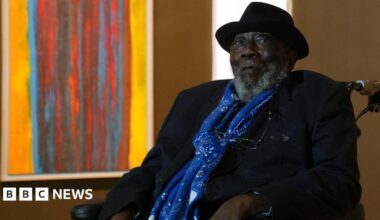 Sir Frank Bowling looks at the camera unsmiling. He is in a wheelchair and behind him is a brightly coloured orange and red abstract painting. Sir Frank is wearing a black hat and jacket and blue scarf.