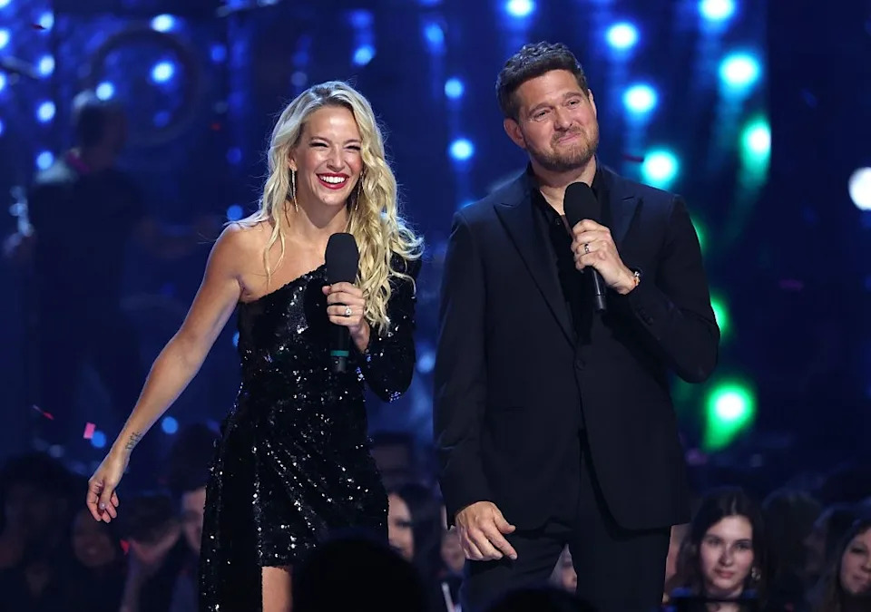 Two celebrities stand on stage, smiling and holding microphones, dressed in elegant black attire at an event