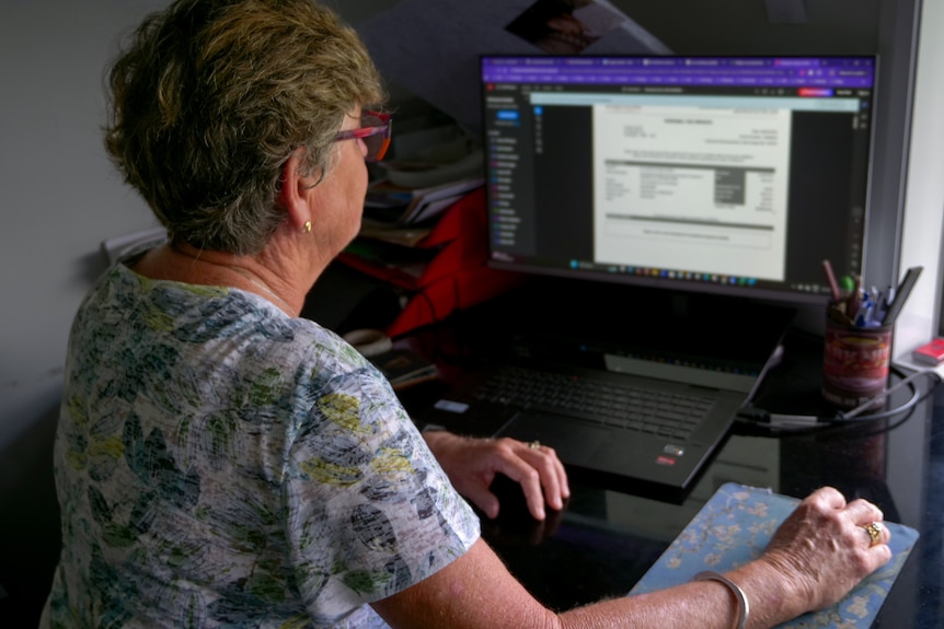 Older woman sits at home office computer with blurred finacial statement on screen