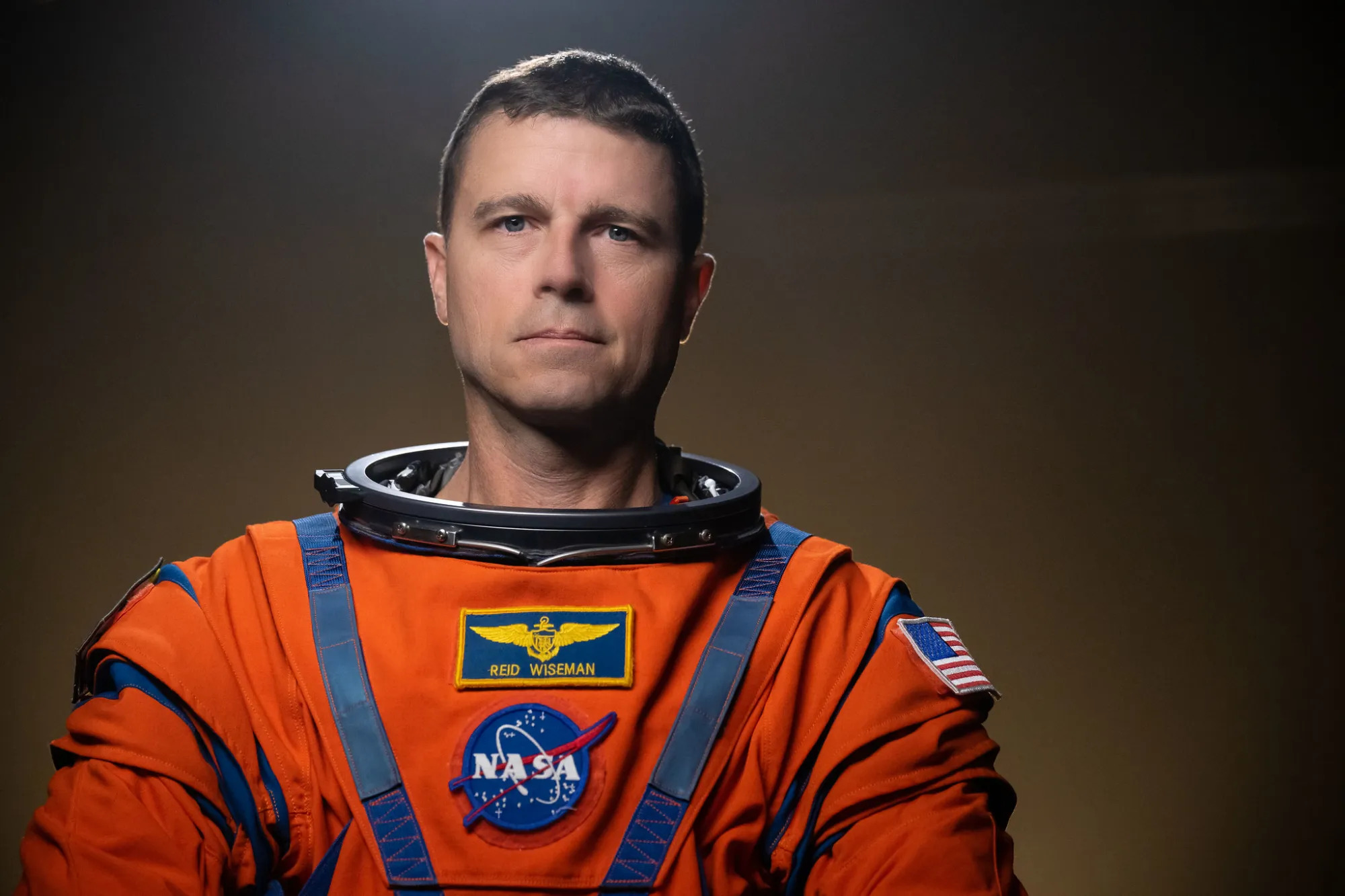 A man with gray hair wears an orange spacesuit in front of a dark background and looks at the camera