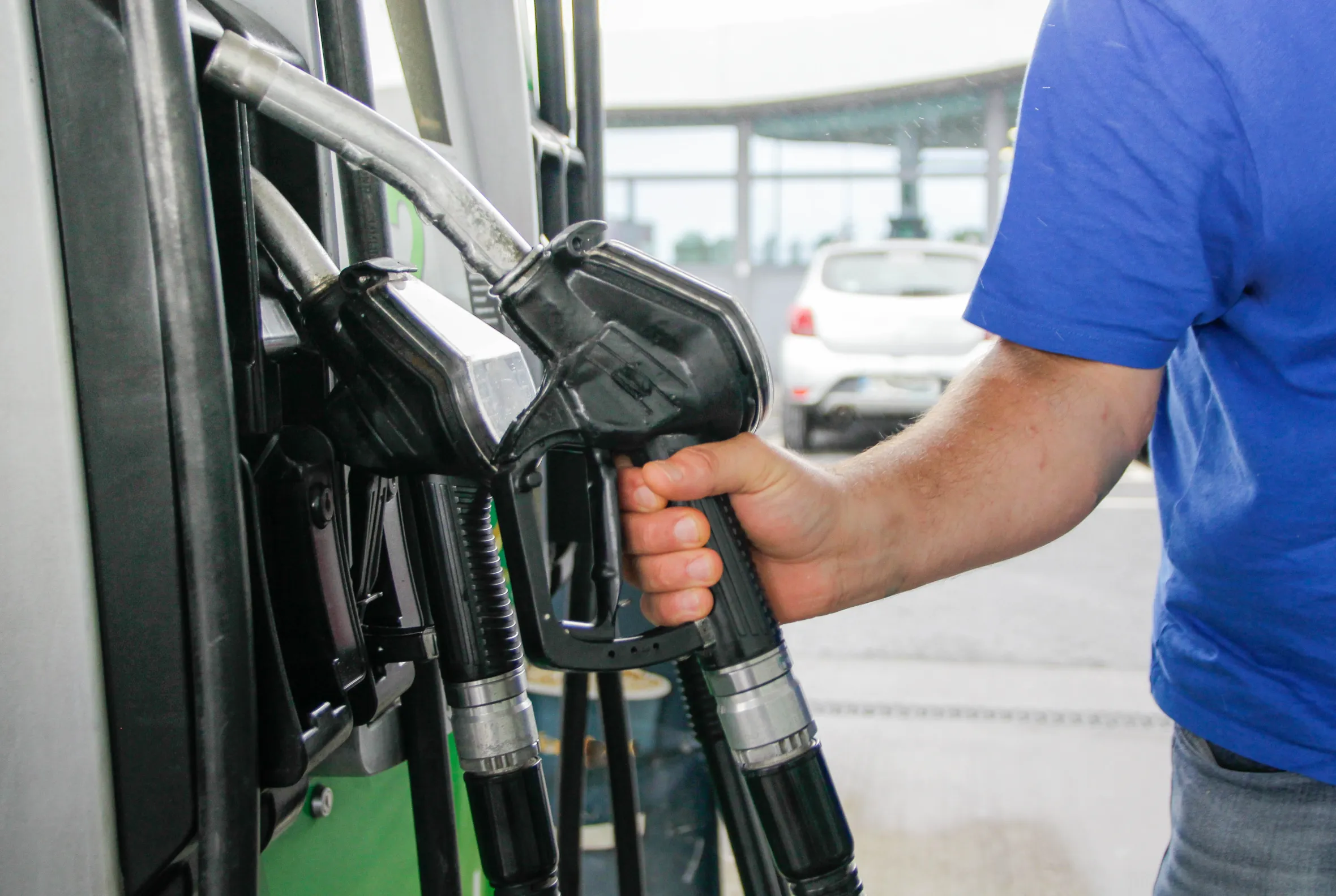 A person in a blue shirt grabbing a gas pump handle at a station with a white car in the background.