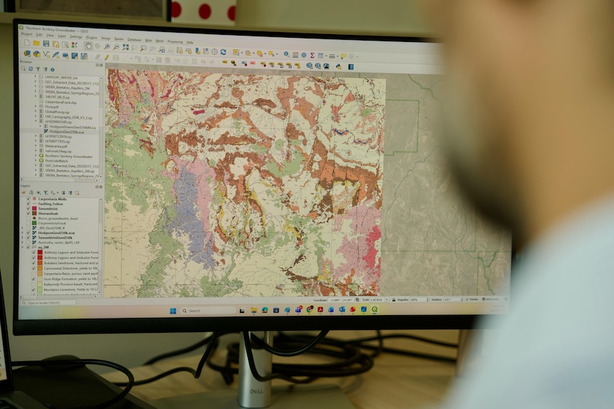 A blurred white man, with beard in foreground, looking at colourful map on computer desktop screen.