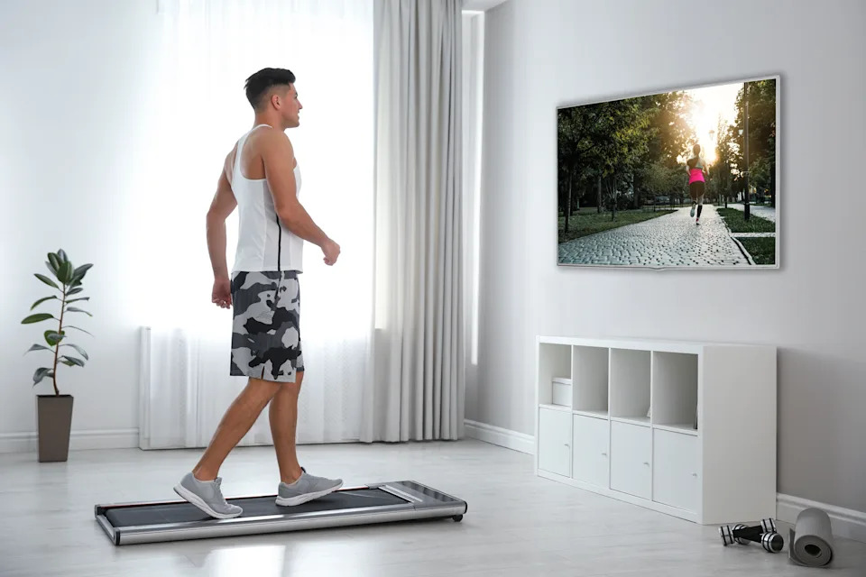 Person walking on a treadmill in a living room, watching a wall-mounted screen displaying a park scene