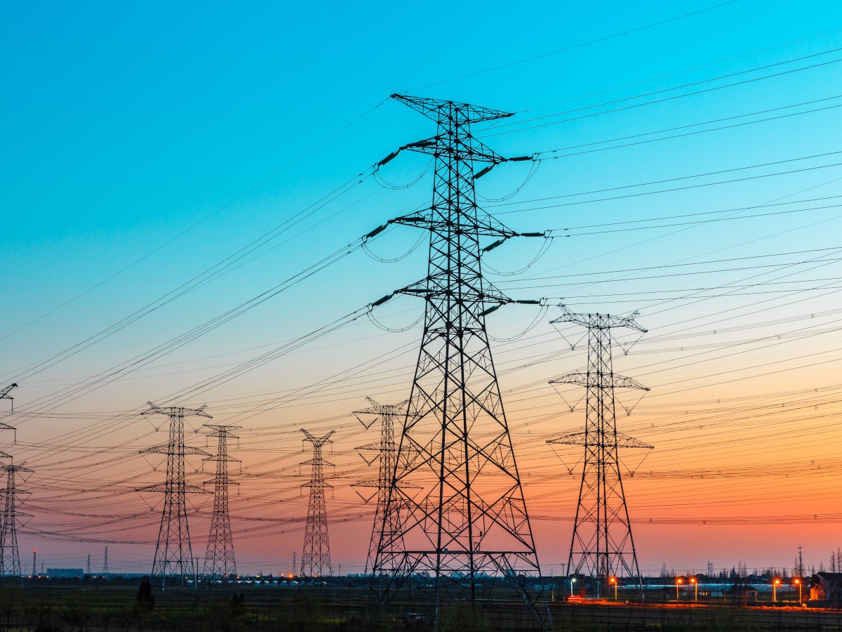 A picture of multiple electricity towers connected by long powerlines. The sunset sky behind them is a mix of blue and orange.