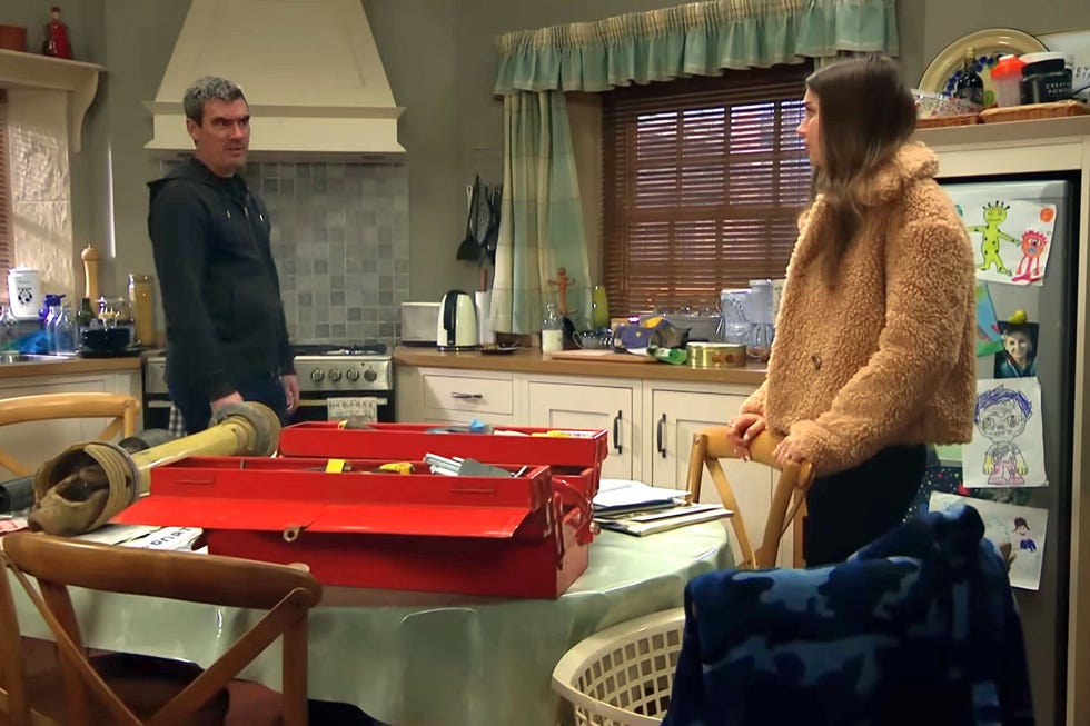 emmerdale, cain and sarah arguing in the kitchen, kain wearing a coat and sarah wearing a fleecy jacket