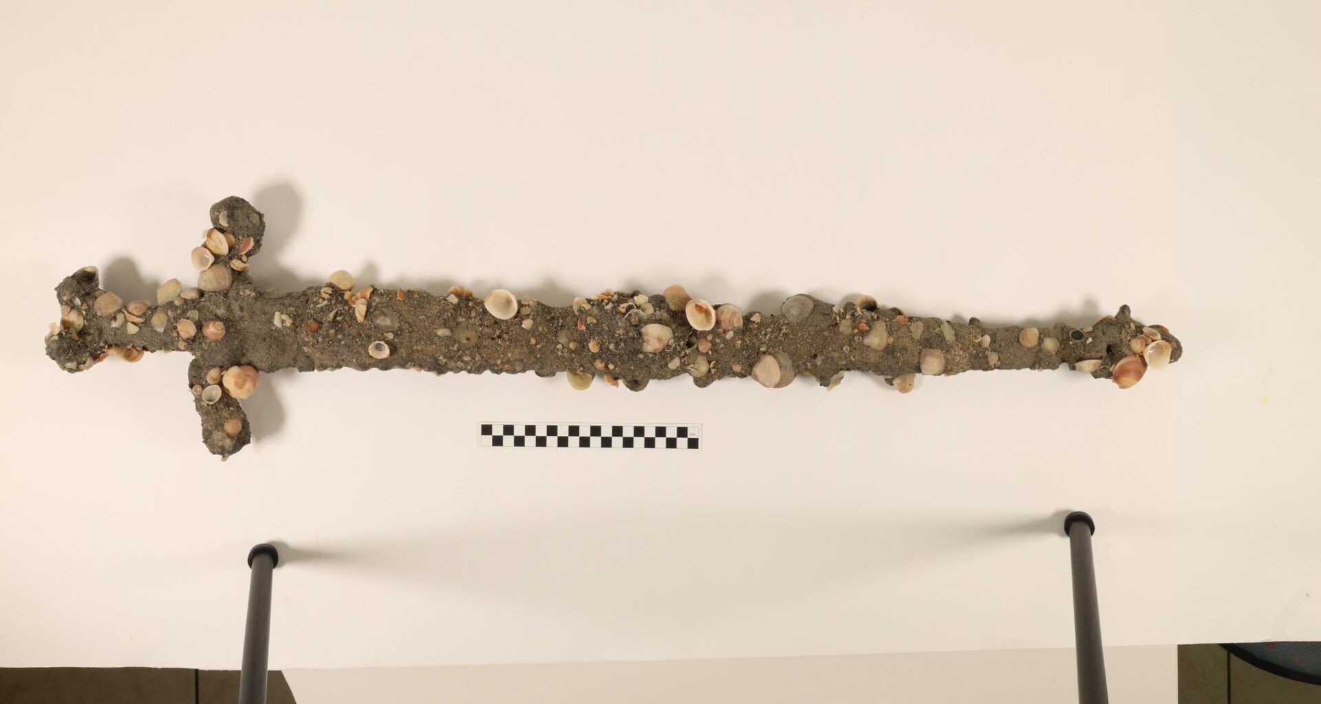 A barnacle and sand encrusted sword hangs on a white wall.