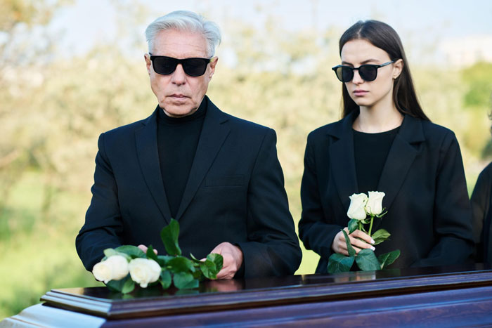 A grieving 27-year-old woman and older man in black at a funeral holding white roses outdoors.