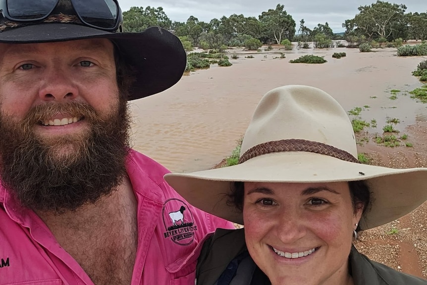 two people with hats on smile at the camera with a body of water flowing behind them