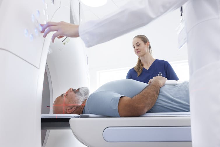 A man lies on his back, entering a CT scanner, with two health professionals overseeing him.