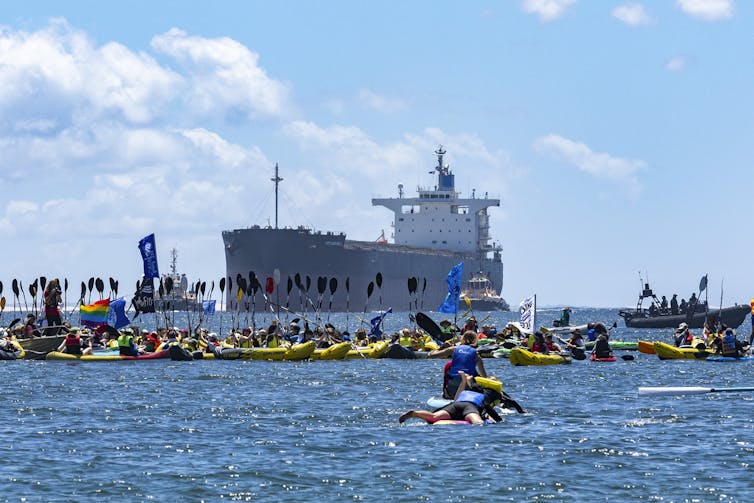 A group of protesters in kayaks paddle along a blue ocean towards a coal tanker.