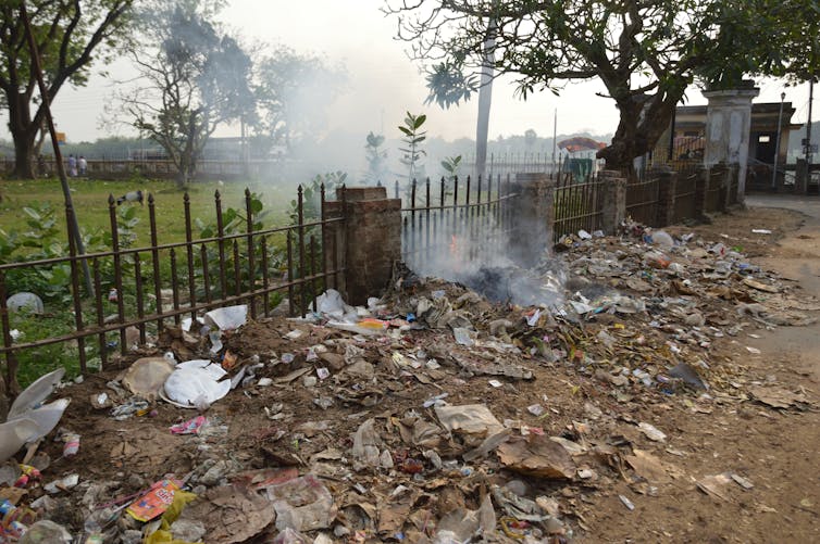 Open burning of mixed roadside waste beside an iron fence, with smoke drifting across a grassy area and trees.