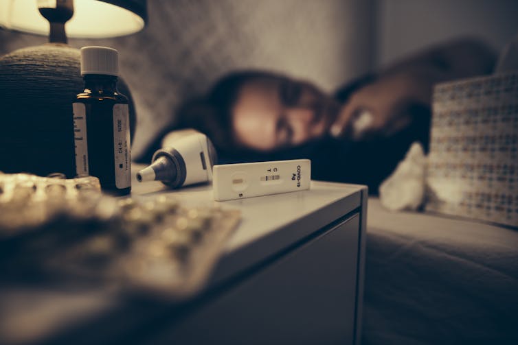Pills, medicin and thermometer and a positive rapid COVID-19 test on a nightstand,with a sleeping person in the background