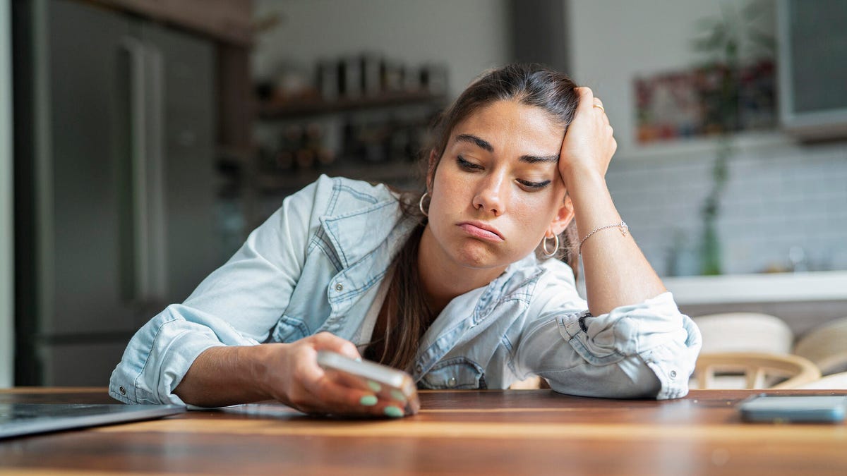 An annoyed woman leans on a table and looks at her phone.