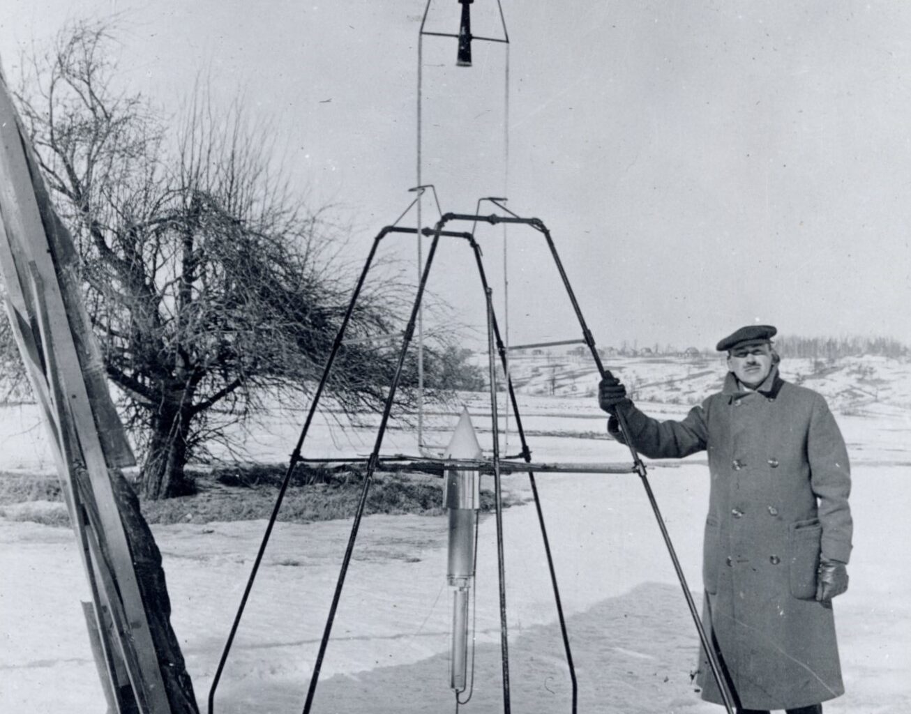 On a snowy March 16, 1926, Dr. Robert H. Goddard rests his hand on the testing frame supporting his liquid fuel rocket at Ward Farm in Auburn, Massachusetts. A wooden door is propped up at an angle next to the frame where Goddard’s assistant, Henry Sachs, later sheltered after lighting the rocket.