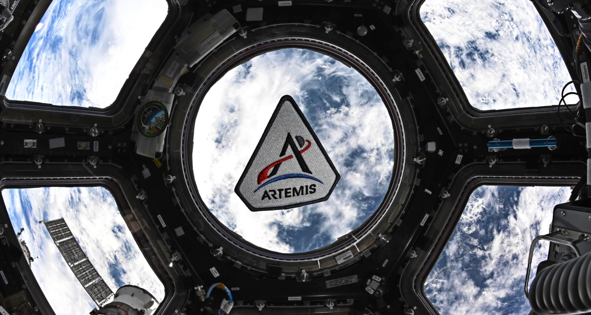 The Artemis program patch in the space station's cupola. The patch is framed by the center circular window in the space. Earth can be seen behind the patch. The Artemis patch has five sides. It has a white background with a black border. There's a black "A," for Artemis. The crossbar of the A is made by a curving red trajectory line that crosses from left to right. That line continues beyond the "A" and beyond a small gray circle representing the Moon. Below the "A" is a blue crescent representing Earth. At the very bottom is the word "Artemis."