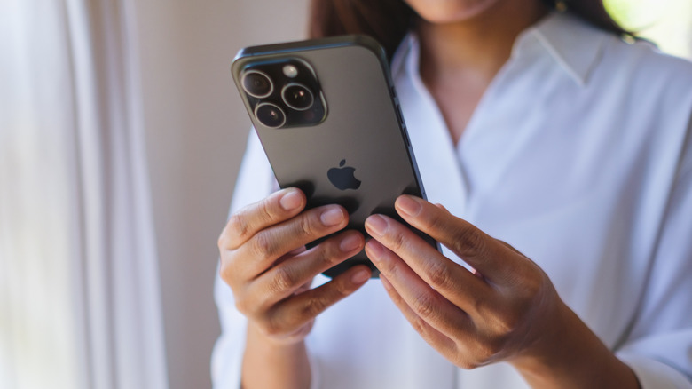 An individual holding a black, 3-camera iPhone model in beytween both of their hands, the individual appears to be indoors and is wearing a dress shirt