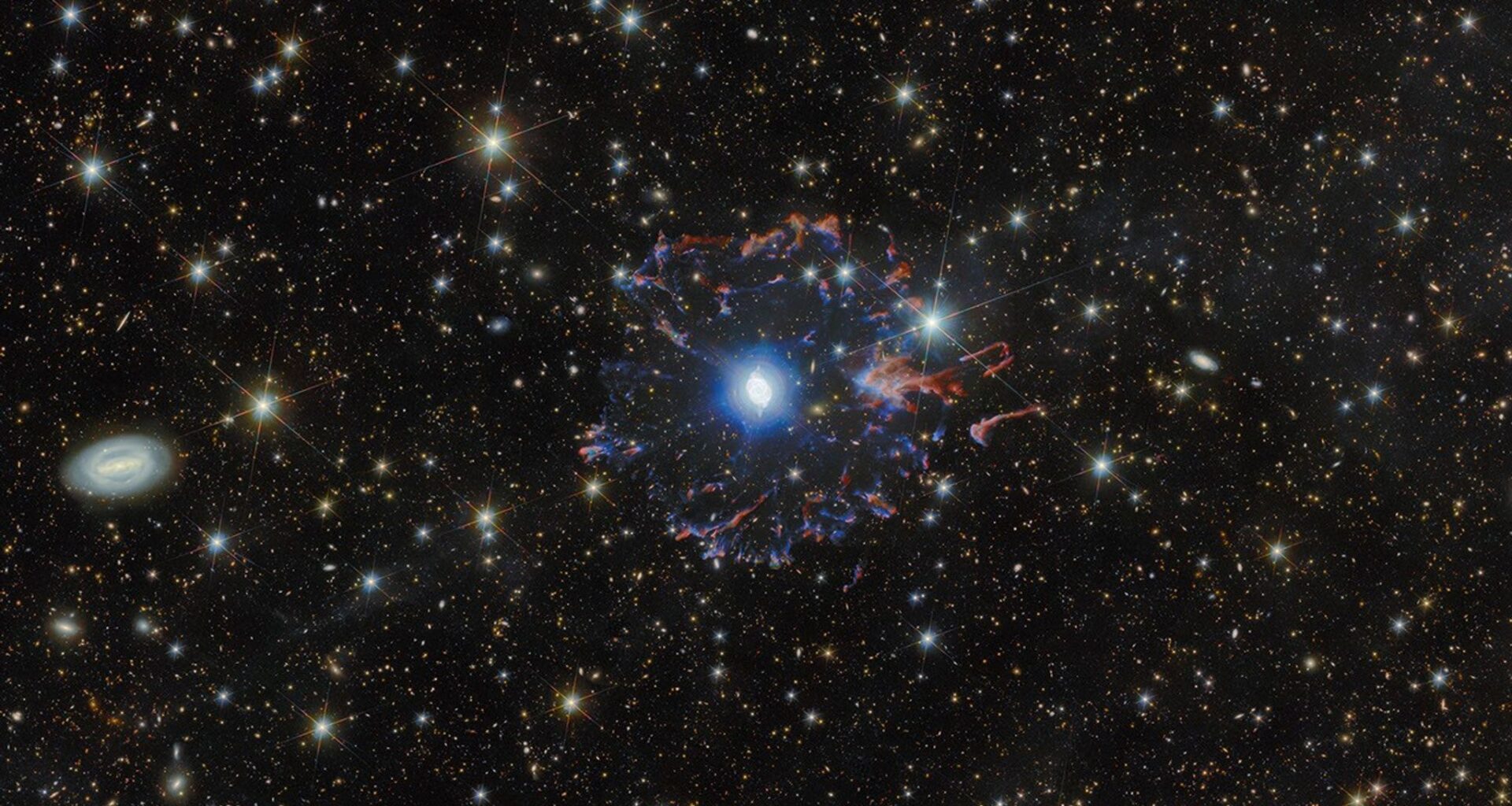 A planetary nebula in space. The star in the very center is surrounded by white bubbles and loops of gas, all shining with a powerful blue light. Farther away a broken ring of red and blue gas clouds surrounds the nebula. A multitude of golden and white stars, wisps of gas and distant galaxies of various sizes surround the nebula on the black background.