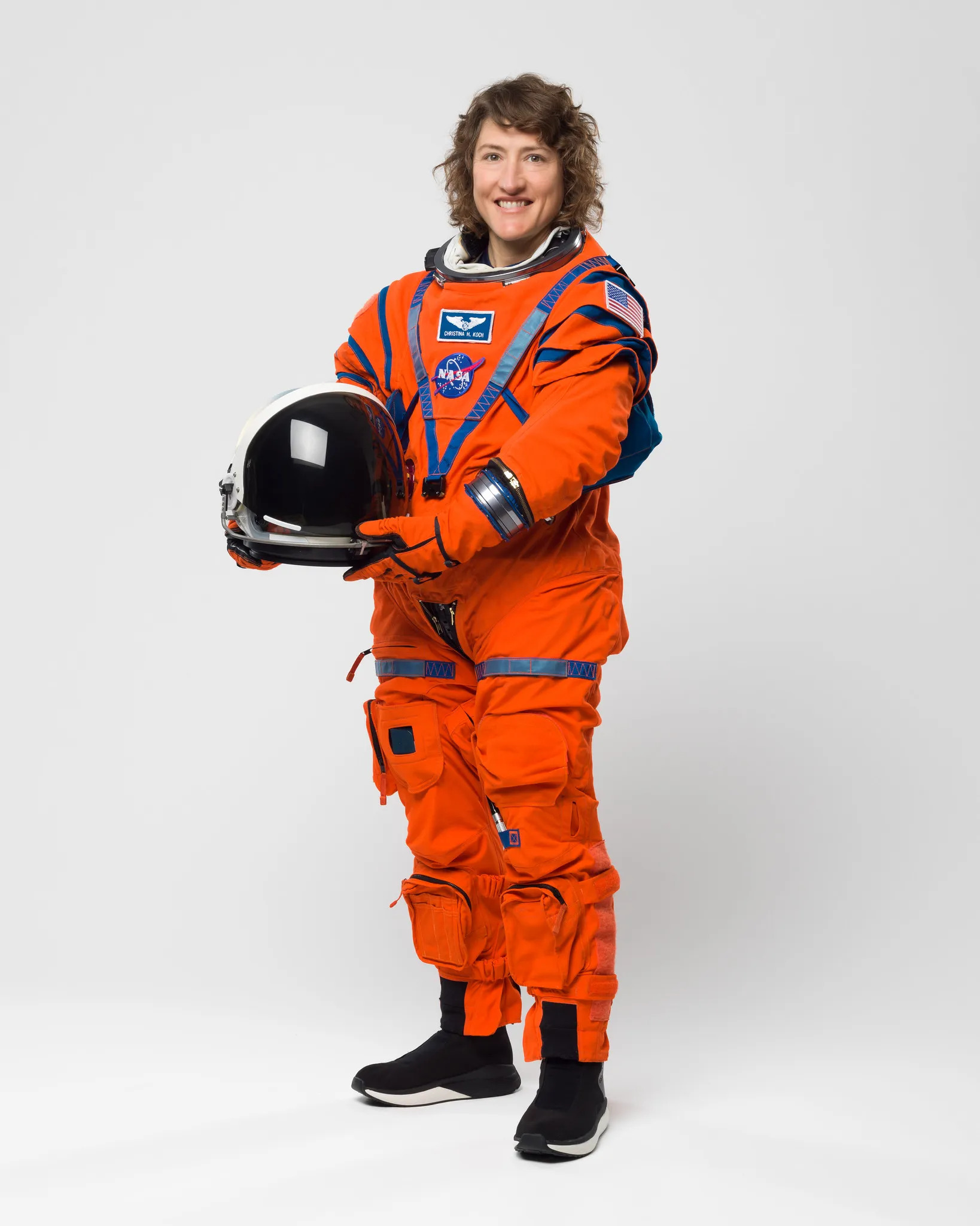 A woman stands in a full profile photo wearing an orange spacesuit and holding a darkened helmet in front of a white background