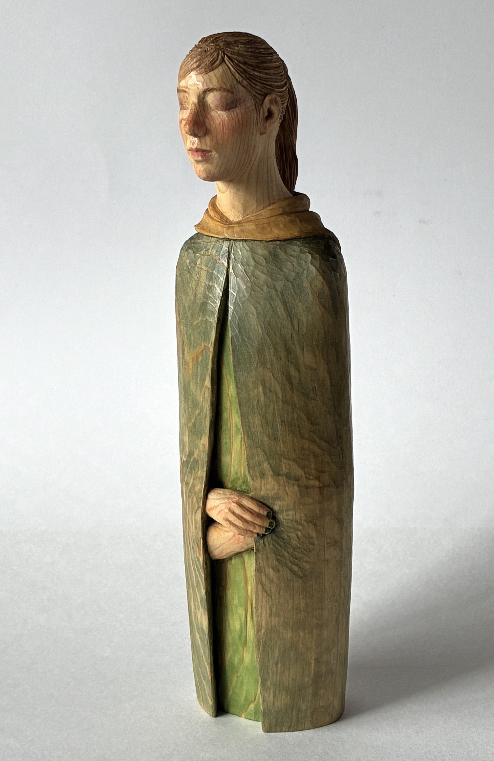A figurative, painted wood sculpture of a young woman in a green cloak, in three-quarter, profile by Shinsuke Inoue