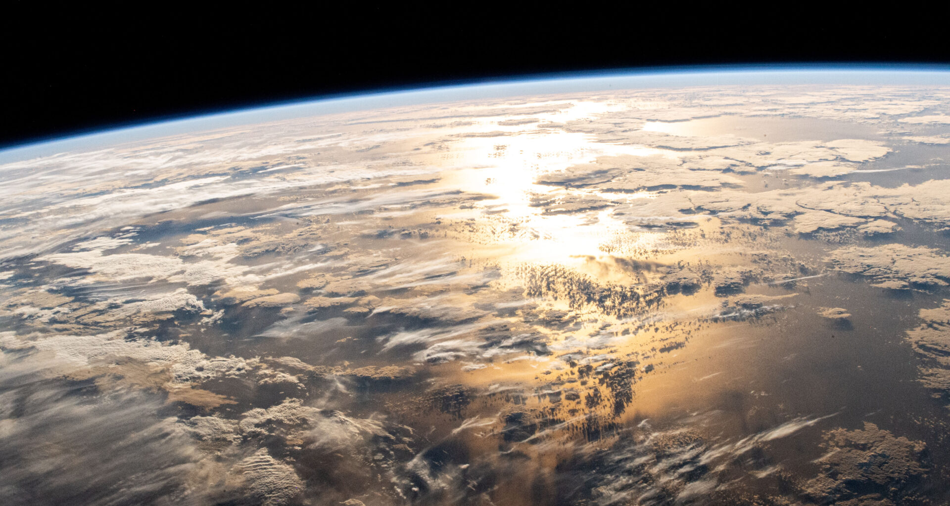 A view of the Atlantic Ocean from the International Space Station. Sun shines on the water, turning the water in the middle of the photo yellow-orange. There are flat, white clouds scattered above the ocean. In the distance, Earth’s gentle curve is outlined by a pale blue hazy line – that’s our atmosphere. Beyond that is the darkness of space.