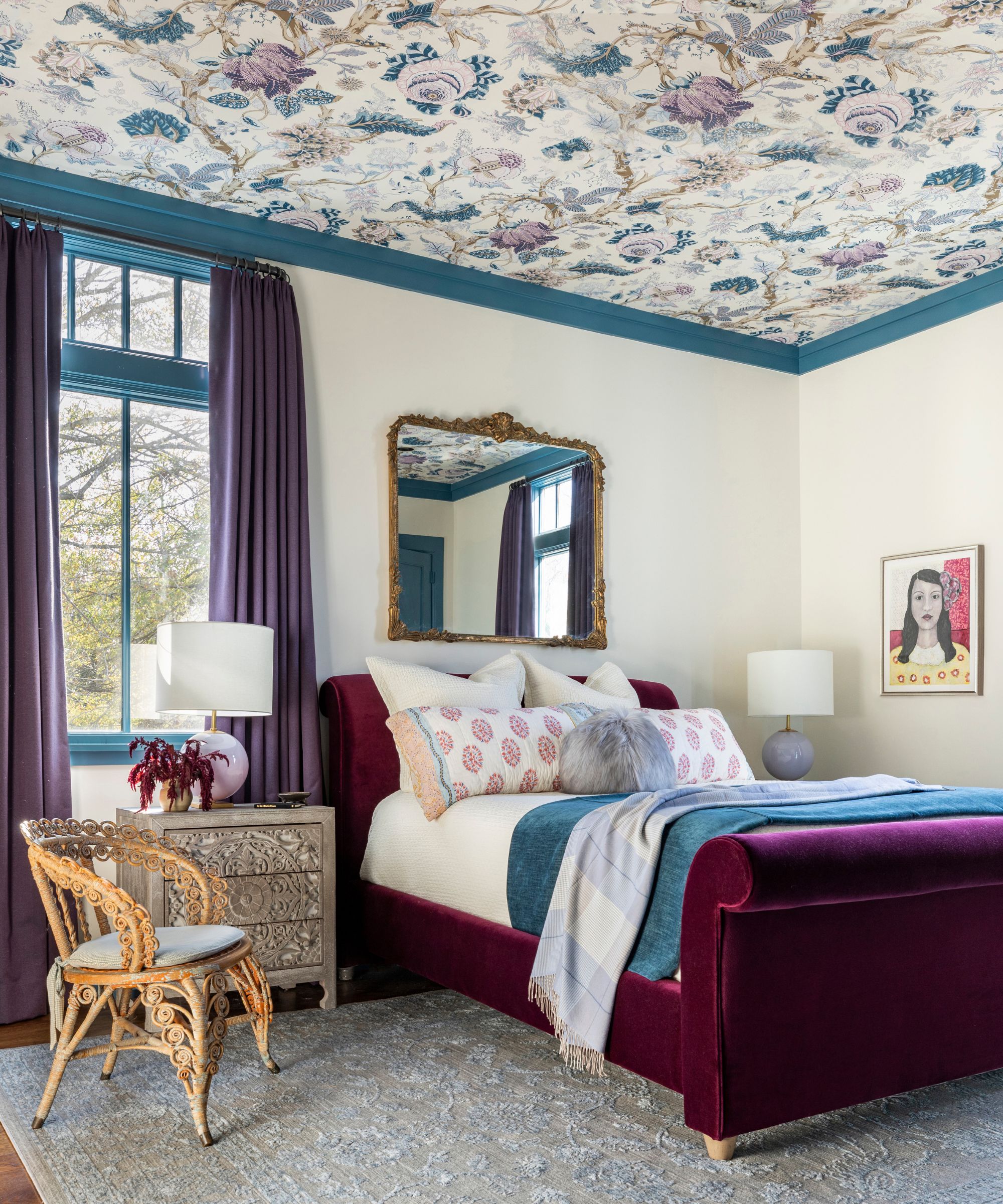 A bedroom with warm white walls, blue trim, and a blue and purple floral wallpaper on the ceiling. Purple curtains and a plum velvet bed.