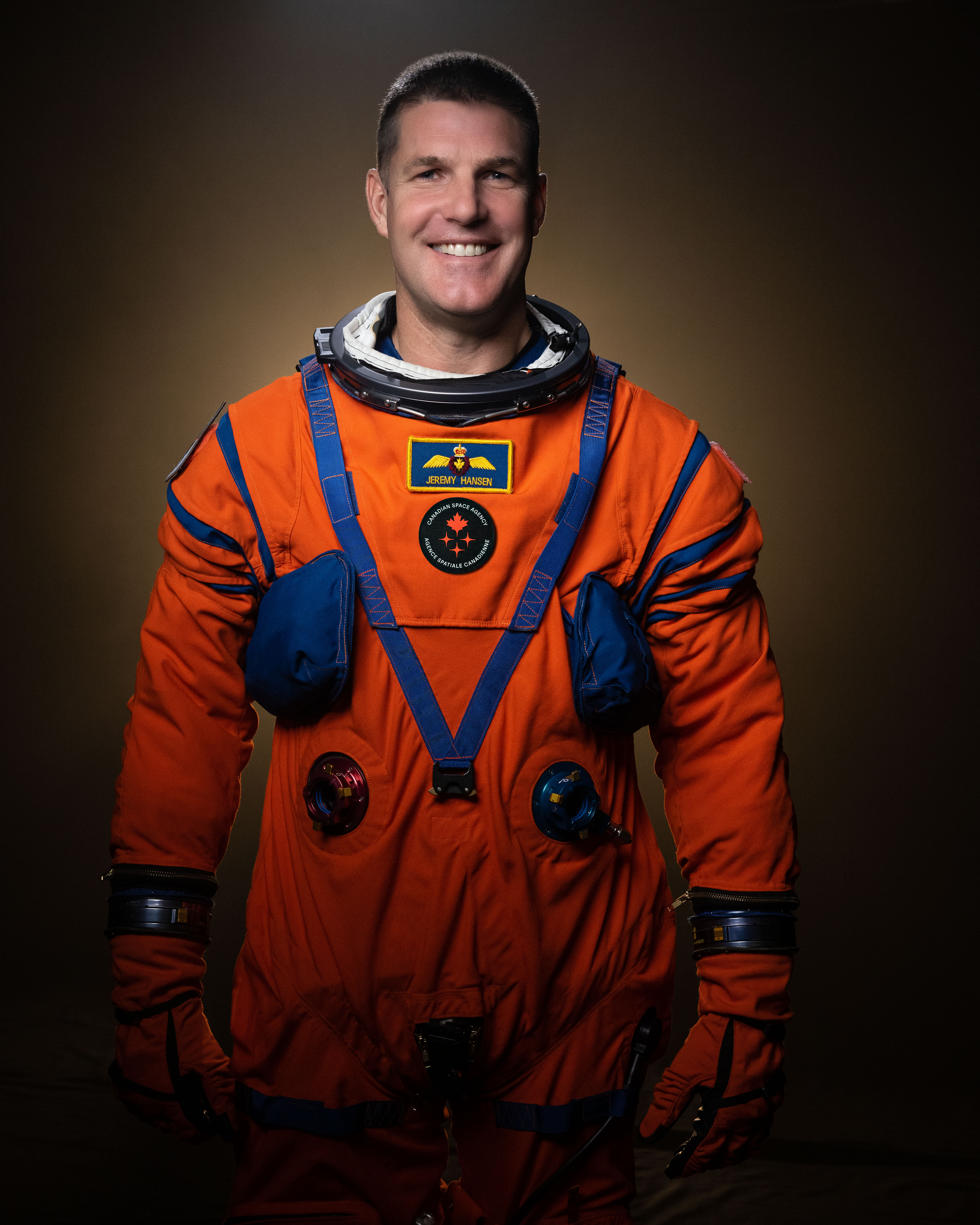 A man with gray hair wears an orange space suit and stands in front of a brown wall