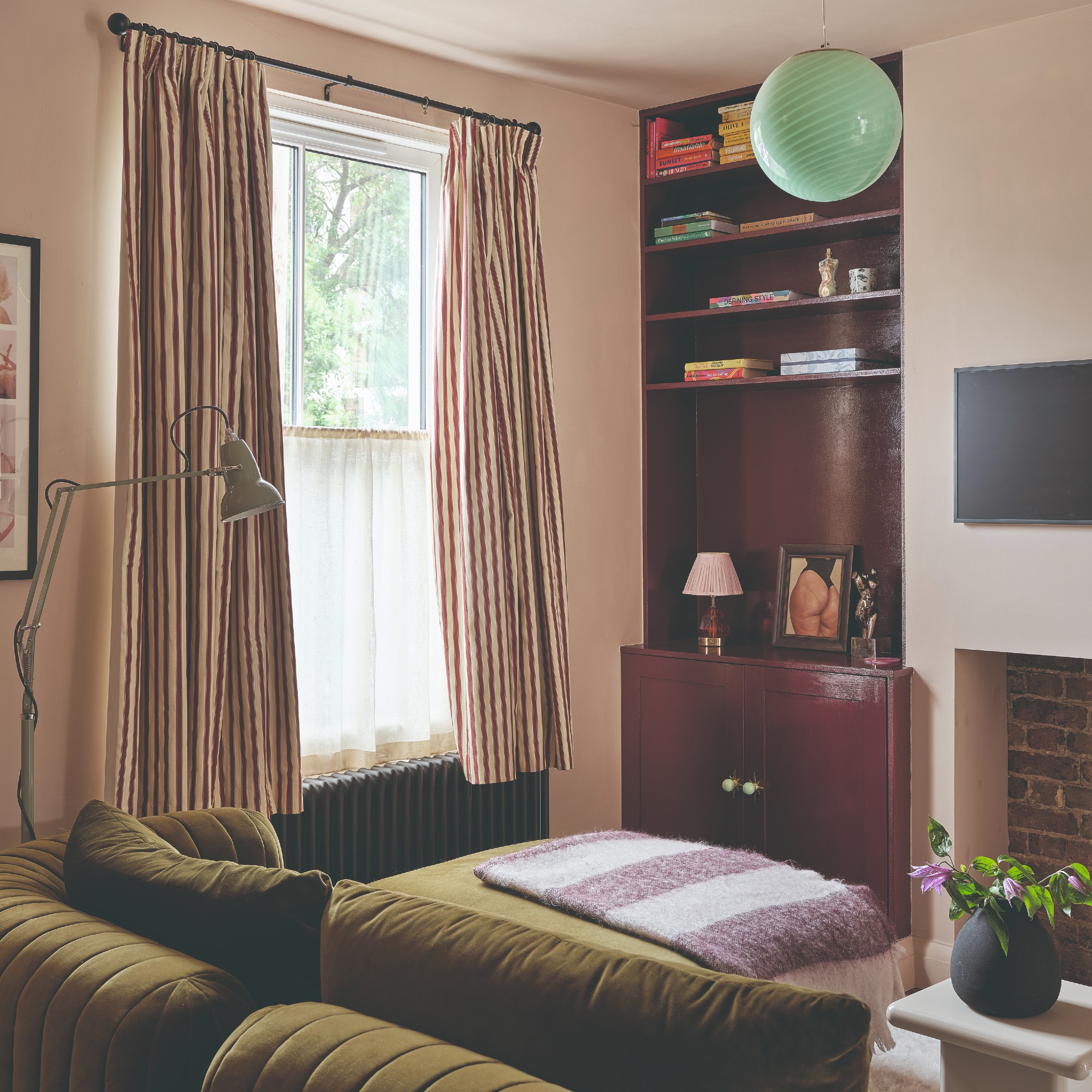 A pink-painted small living room with a green velvet sofa and striped short curtains on the window
