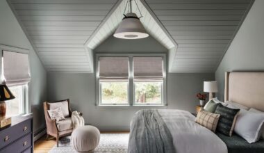 A bedroom with color-drenched pale green walls and a sloped ceiling with panelling. A large taupe headboard and layered neutral-colored bedding, cushions, and throws.