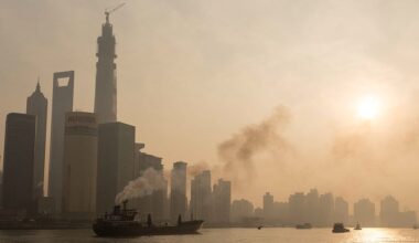 A smog-filled cityscape on the edge of a body of water is seen under a clouded over sun