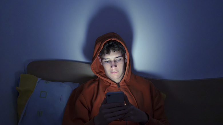 Young man in hooded sweatshirt in bed looking at phone in a dark room