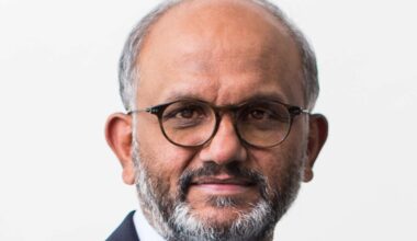 A man with a grey beard and glasses smiles at the camera wearing a suit.
