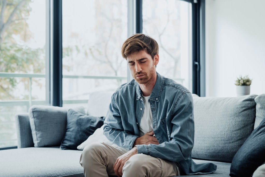 A young man sits on a couch, holding his stomach in pain.