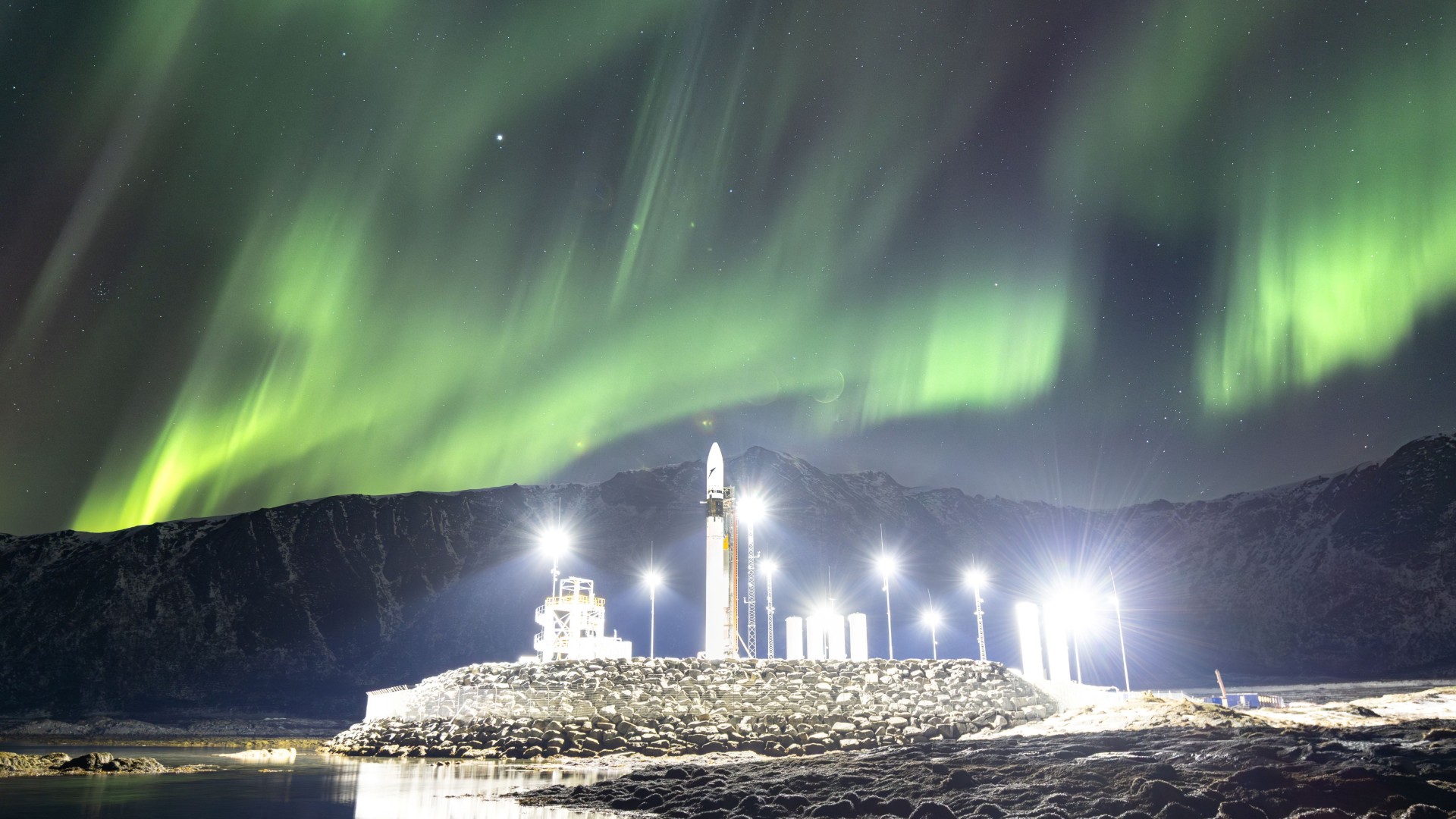 a white rocket stands upright on a launch pad surrounded by lights, while green wisps of light streak across the starry night sky overhead. the silhouette of a snowy mountain range looms in the background