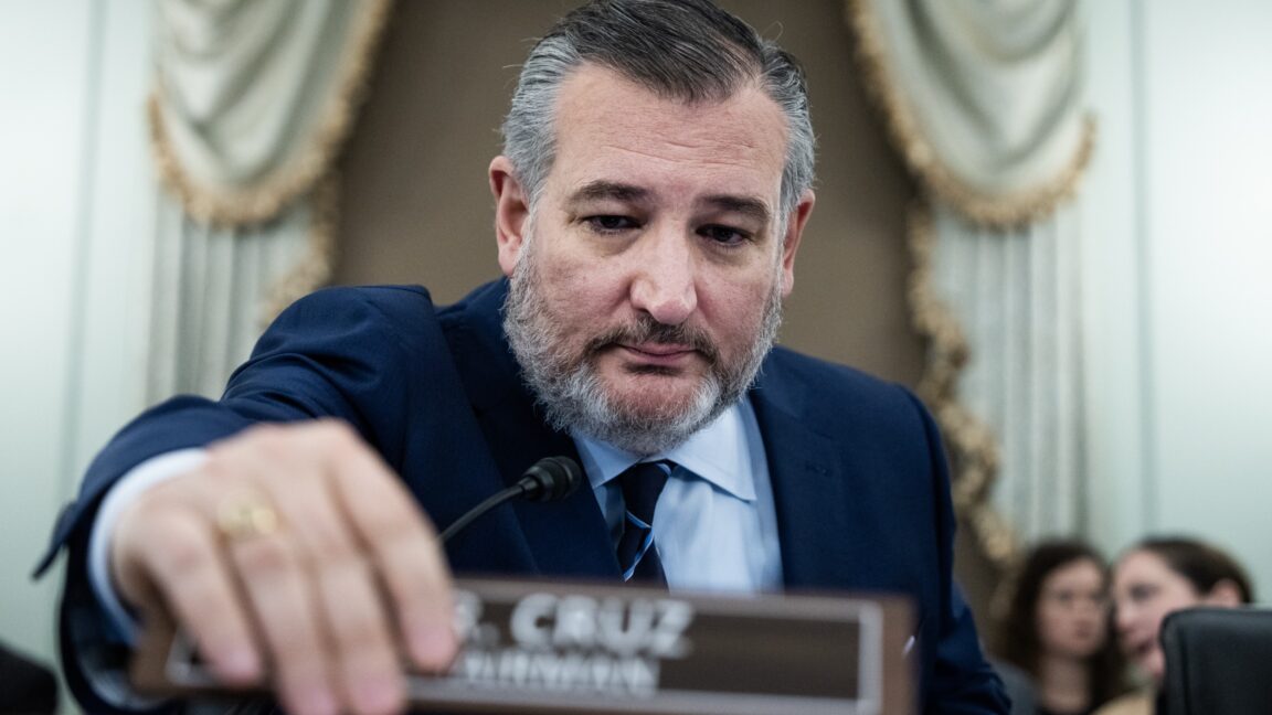 Senator Ted Cruz at a Senate committee hearing, sitting in his seat and using his hand to move a nameplate that says "Mr. Cruz, Chairman."