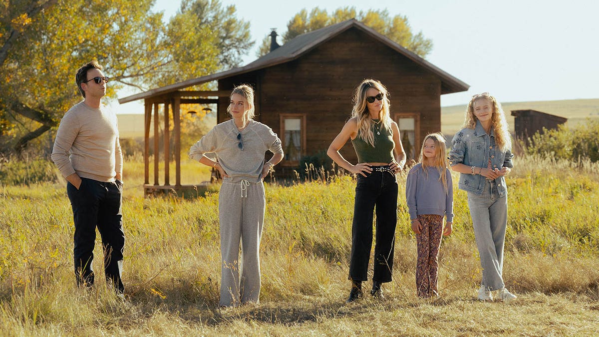 Patrick J. Adams as Russell McIntosh, Elle Chapman as Paige McIntosh, Beau Garrett as Abigail Reese, Alaina Pollack as Macy Reese, and Amiah Miller as Brigitte Reese standing in tall grass in front of a rustic wooden cabin on a sunny day.