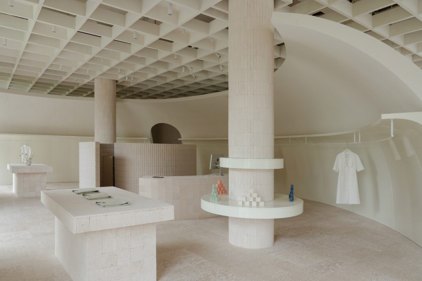 Image of a minimalist, neutral retail interior made from limestone and travertine. A truncated dome partition wall encloses the space, with two structural columns, two stone tables and display shelves.