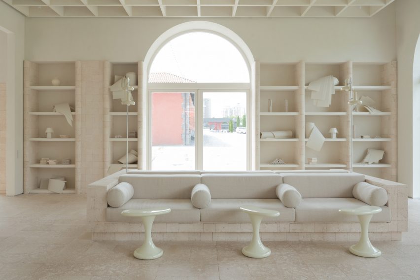 Image of a low-level boxy sofa, with small organic-formed side tables and tubular cushions. Built-in shelving units can be seen on the back wall, separated by a large arched glass doorway.