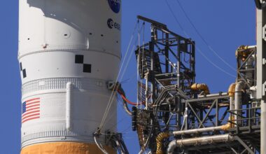 A section of a rocket seen with multiple lines, or umbilicals, connecting it to eqiupment on a tower.
