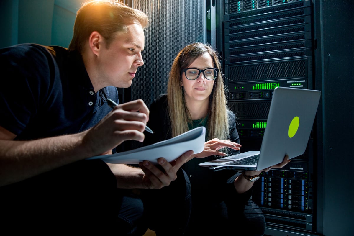Two engineers, one writing in a notebook and the other using a laptop, are working in an AI data center.