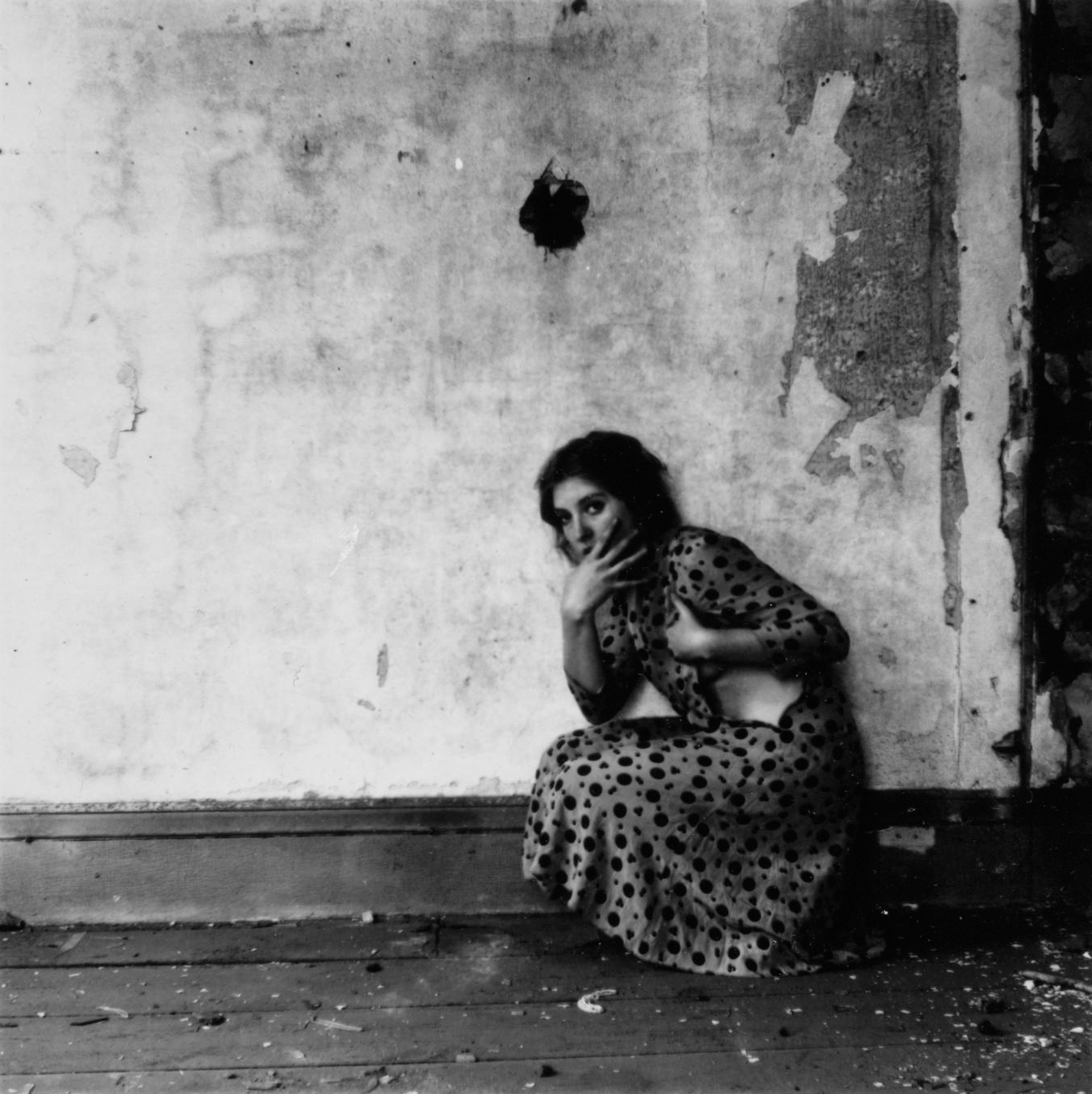 A woman in a polka dot dress crouches in a corner of a decaying room with a distressed expression