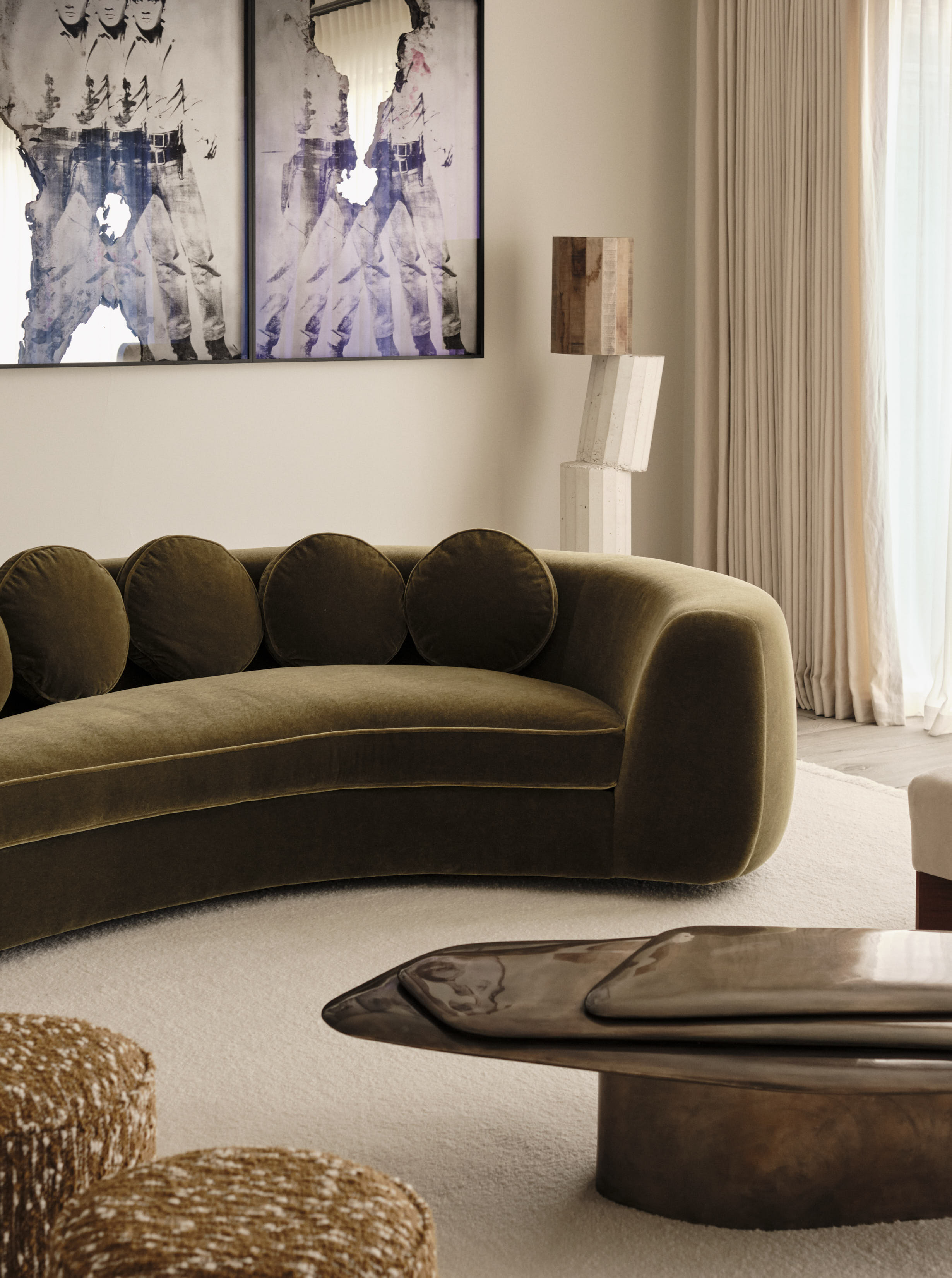 modern living room with green velvet curved sofa with sculpture behind, framed artwork, coffee table, beige rug, and beige curtains