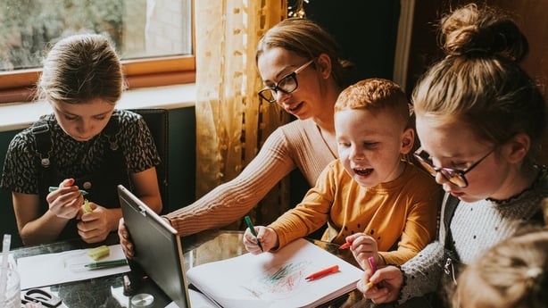 Single Mum in a home environment home schools / helps her children with homework. She holds a digital tablet out in front of her. Recognisable scene for parents in lockdown attempting to juggle a work / life balance during the Coronavirus pandemic.