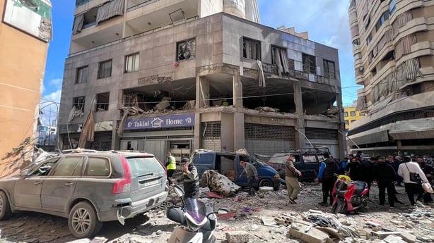 People walk among debris and rubble with damaged cars and buildings