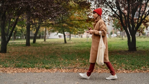 Female in warm coat and knitted hat holding smart phone and walking in park.