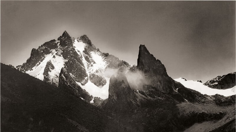 Snow-capped mountain peaks with rugged, jagged edges rise sharply against a cloudy sky. Mist and clouds drift around the rocky summits, highlighting the dramatic, contrasting landscape.