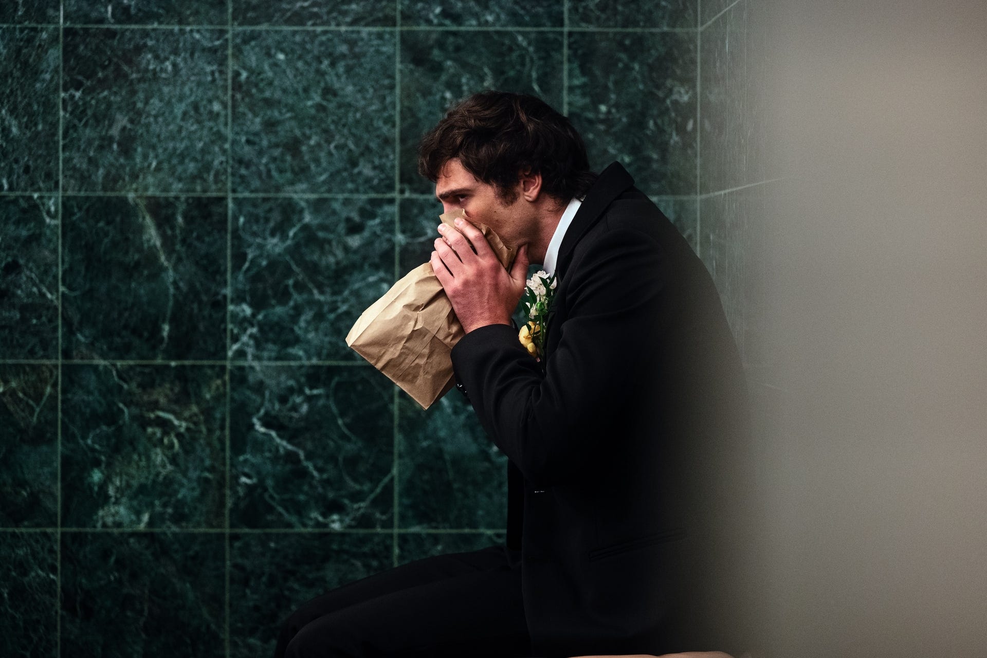 A man in formal attire sitting in a restroom with a paper bag.